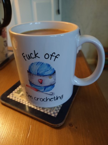 Mug of tea on a bedside cabinet, it says 'fuck off I'm crocheting' in the centre is a ball of blue yarn with a crochet hook 