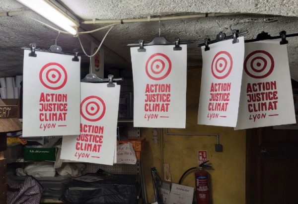 Accrochées au plafond d'une cave, 5 affichettes "Action Justice Climat Lyon" ornées du logo, 3 cercles concentriques légèrement irréguliers (le plus petit est plus un point qu'un cercle)
