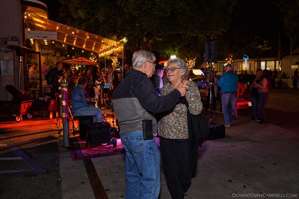 A couple of seniors locked hand in hand on a darkened street, dance on a sidewalk to a band playing underneath the orange glow of dim outdoor mood lighting in the background.