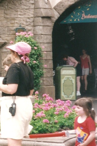 People at a theme park, with a woman in a black top and pink hat, a child in a red shirt, and others in the background near a building with a light green trash can. There are pink flowers in the foreground.