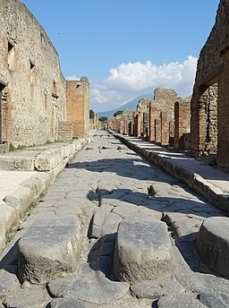 A Roman street in Pompeii.  Looking down a stone-paved road between ruined buildings. © Ad Meskens / Wikimedia Commons