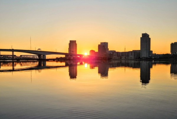 The rising sun has emerged from behind the city skyline silhouettes of buildings and bridges over the calm Saint Johns River. Sending reflections upon the surface like a reverse mirror and casting an orange glow over the horizon.