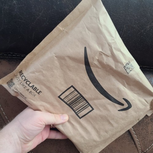 Photograph of a white person's hand holding a brown, Amazon package with what looks like books inside against a brown couch.