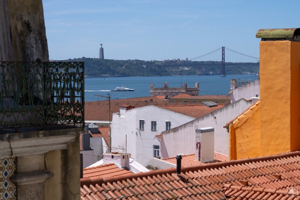 Color photo of white and orange buildings with red tile rooftops with Lisbon’s Tagus River and a large suspension bridge spanning it beyond them. 