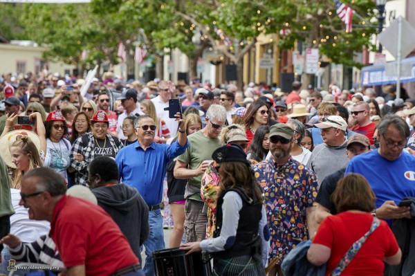 Photo of a large crowd of people walking in all directions down a closed street after a 4th of July Parade. Lots of people are wearing festive red, white, and blue accessories. Slightly off center is one very noticeable person in sunglasses taking selfie.