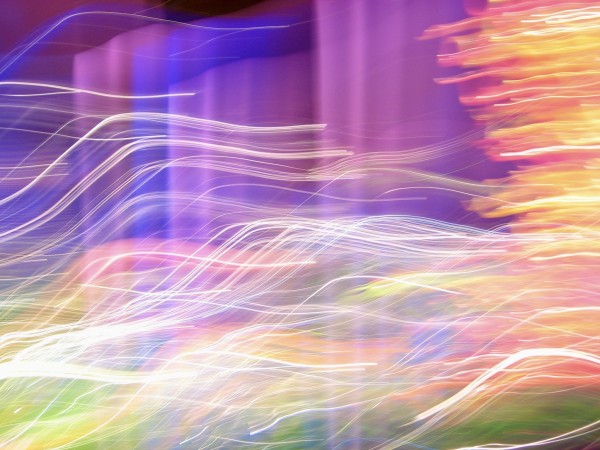 Wiggly lines of white and other light colors, yellow green and amber and blue in horizontal waves, against a purple and pinkish vertical background. Long exposure moving-the-camera shot of a colored glass art exhibit creating the lines and illusion of movement.