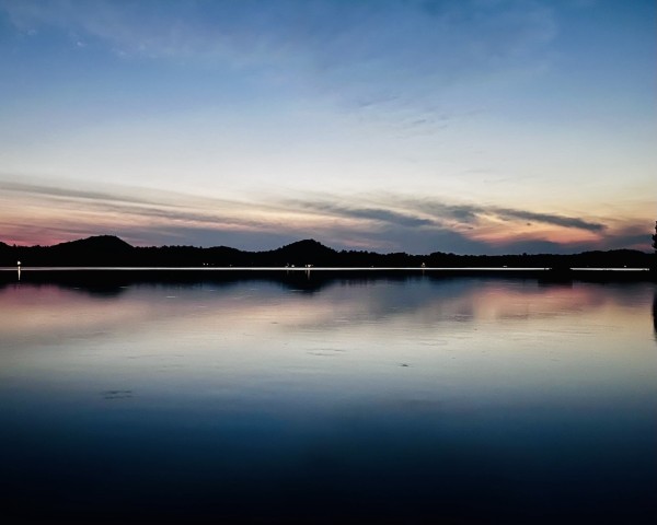 Twilight landscape showing a calm lake with cloud reflections which look like the feathered tail of an arrow, silhouetted hills on the horizon, and a colorful sky.