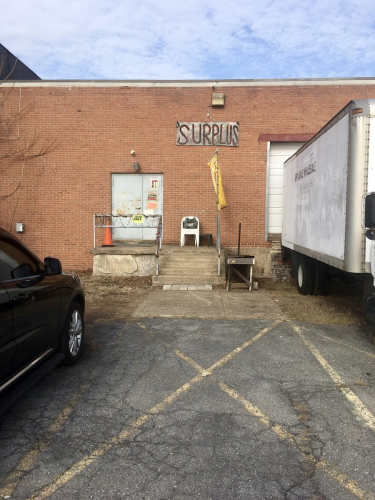 An industrial flat-roof brick building with a loading dock and stairs up to two metal doors. A sign says "SURPLUS", written in Charlie Brown-like letters with characters decreasing in width as they go left to right.