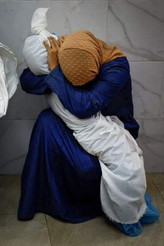 A woman in Gaza, in a blue dress wearing a yellow headscarf, crouched in a corner of a building. Her arms are wrapped around the dead, tiny body of a little girl wrapped in white cloth. The woman is covering her face in her own arms, mourning.