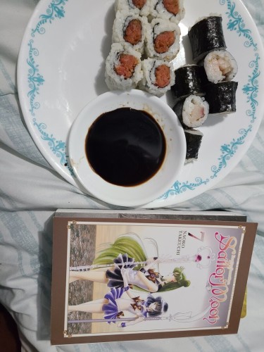 Sideways photograph of Volume 7 of the Naoko Takeuchi Collection edition of Pretty Guardian Sailor Moon sitting on a white / blue comforter with a plate covered in sushi on it. Six of the pieces have a pinkish fish in the center and rice on the outside; six of the pieces have a white fish in the center with the seaweed on the outside.