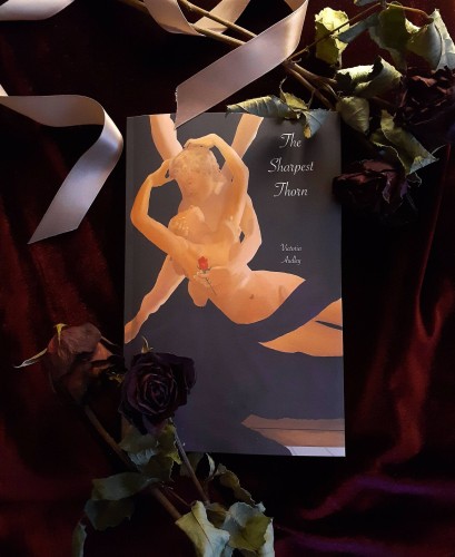 a paperback copy of the sharpest thorn, which features on its cover a replica of the antonio canova statue of eros and psyche embracing. psyche is draped with purple fabric and eros holds a red rose in his hand. white cursive text reads: the sharpest thorn, victoria audley. the book sits on a background of dark red velvet, and is surrounded by a pale purple ribbon and dried dead red roses.