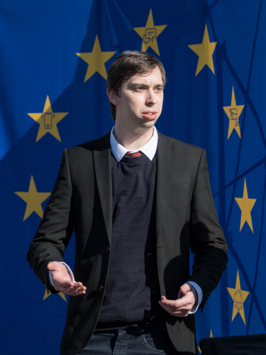 A person speaking in formal attire in front of an EU flag. At closer look, some stars in the EU flag contain icons like a CCTV camera or a smartphone.