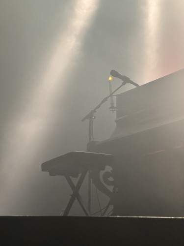 A piano and microphone stand illuminated by stage lighting and a lit candle, with atmospheric haze.