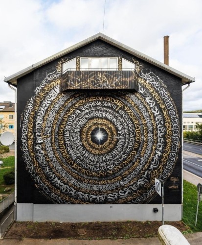 Streetartwall. The impressive mural with circles of calligraphy lettering was sprayed/painted on the outside wall of a multi-storey building with a balcony in the gable. The building has a short, narrow chimney. The large wall is painted in black, gray, silver and gold. On a black background, 13 rings of tattoo-style lettering are painted around a bright atrium. The colors and sizes of the lettering change depending on the circle. Even the lower part of the balcony has been included. (The photo shows a narrow brown lawn in front of the house, adjoined by a street and houses).
