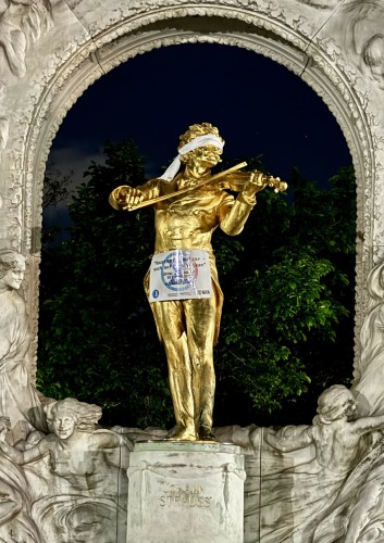 A night shot of the golden statue of Johann Strauss playing the violin, blindfolded.

A leaflet in German is stuck to his body:

"The main thing is to waltz - even on a volcano.

Open your eyes, be honest.

We are in a climate emergency".

Photo credit: Extinction Rebellion
