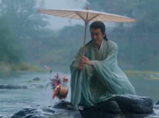 A man wearing traditional hanfu clothes sitting on a rock by a river, holding an umbrella, with a colorful bird beside him. 