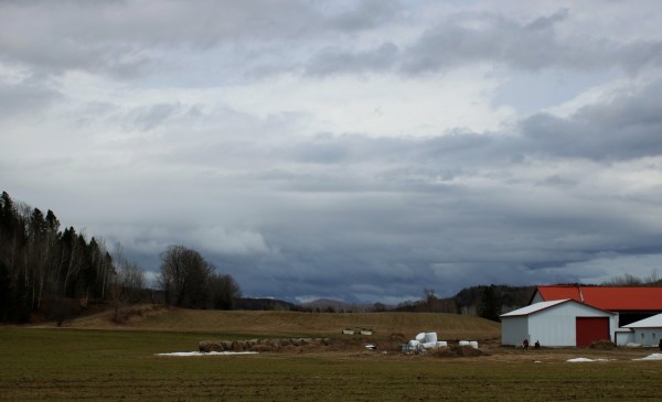 Photography of a sky darkening with grey clouds gathering and seeming to move fast, over a yellow and green field with red and white farm buildings.