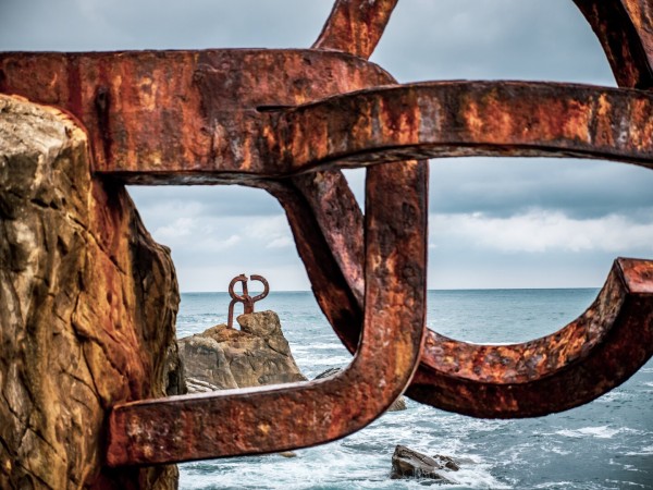Metal structures built into rocks right at the sea, with waves below them. The metal art is rusty with the red rust complimenting the blue water of the sea.
