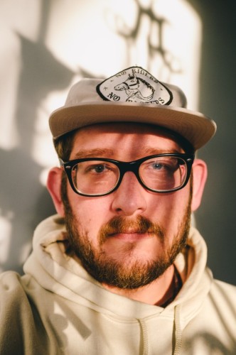 A self portrait taken of a bearded man with black rimmed glasses wearing a stone colored baseball cap and tan hoodie. The  subject is standing in front of a window that is being bathed in sunlight. The sunlight from outside is casting shadows on the wall behind the photographer and shadows on the photographers face.