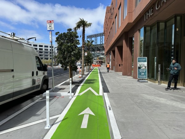 A green, sidewalk-level bike lane on Third Street looking north towards Oracle Park, with a red brick building on the right