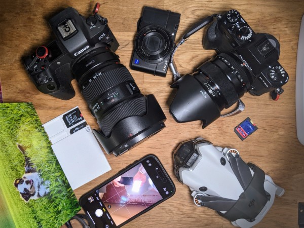 left to right: eos r+adapter+ef 24-70 lens; damaged old rx100ii; x-t30 with a 24mm lens and lenshood, below a print of a dog playing in grassy farmland, sdcards, iphone that reveals someone holding a phone and a light for the picture here; a dji drone and another sdcard 