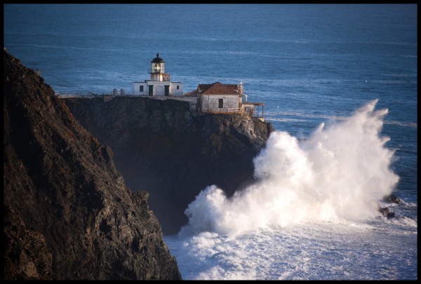 A viee of a lighthouse and utility building on a rocky cliff overlooking the Pacific Ocean. 

The buildings are dirty white, with red roofs. The ocean is very blue. And most significantly, the cliff is being smacked by a giant 40-foot wave.