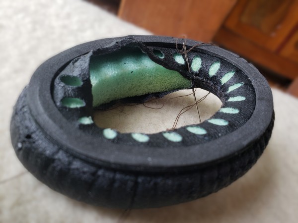 A black rubber headphone cushion is ripped in the centre. Part has been stichted together. Another part is still separated. A needle is sticking into the material to show what is going on.
