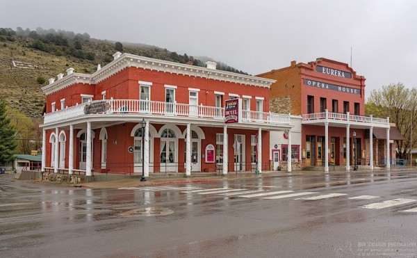 A color landscape photo of two late 19th century far west Victorian style buildings. Both are made of brick with white painted trim. Both have second story walk around balconies. The slightly short hotel is nearest the camera with the slightly taller opera house further down the street to the right. A small one story building separates the two larger buildings. A light green hillside is in the background. It is a rainy day with low clouds and wet pavement. 