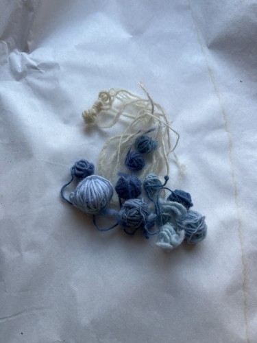 Several small rolled up balls of hand spun wool string, all but one dyed a shade of blue. The shades range from dark ultramarine to periwinkle. 
