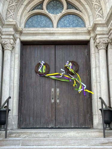 The wooden door of an old church. On the door is a wicker wreath with long white, yellow, and purple ribbons blowing in the wind. The church is made of brick and stone.