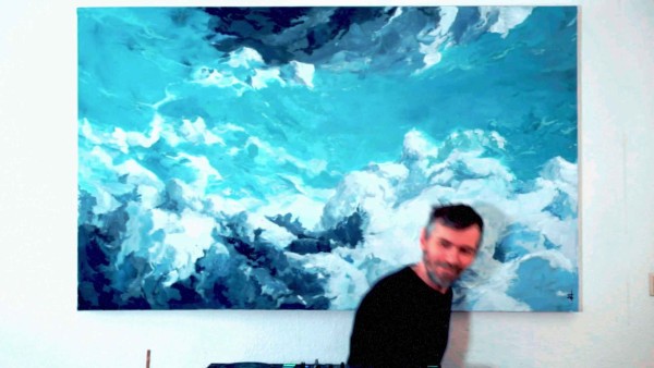 The image shows an individual standing in front of a large canvas with a painting that has a turbulent, abstract motif resembling waves or clouds in various shades of blue and white. The figure of the individual is blurred, suggesting movement or a low shutter speed during the capture of the image. There's equipment on a surface in front of the individual that could be related to music production, considering the context and the image quality, it’s difficult to make out the details. The painting has a dynamic and somewhat stormy aesthetic, and it's the most striking element in the photograph due to its clarity and vibrant color contrast with the surrounding elements.