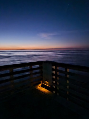 Still quite dark during the "blue hour" prior to sunrise, a view over the vast Atlantic as far as the eye can see, from the corner of an old wooden pier. There's a small orange light illuminating the pier, at the base of the corner. Centered over the water, the corner points to the horizon now colorful with layers of yellow and orange and blue.