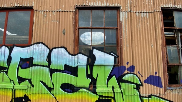This image shows a section of a building with a rusted corrugated metal exterior and two large, worn windows. The metal surface is adorned with a striking graffiti piece featuring black outlines filled with green, white, and purple. The graffiti incorporates a white circle that aligns with one of the windows, creating an optical illusion. The overall scene contrasts the dilapidated industrial condition of the building with the vivid, contemporary artwork.