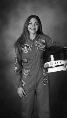 Picture of a very happy 19 yr old female with long hair dressed in a NASA uniform. She holds her enormous helmet on her hip. She's going to Mars!