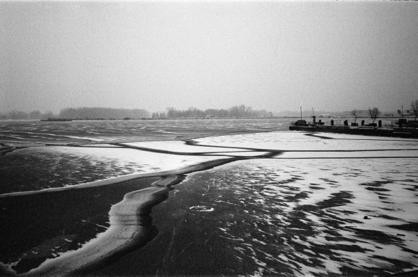 A black and white photograph of Toronto Harbour in winter. The wind is whipping snow across the thin ice formed between piers.