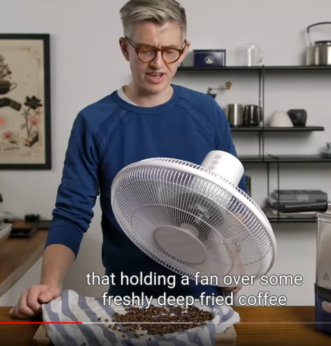 A picture of a guy holding up a fan against coffee beans that have been deep fried, in order to cool them down.