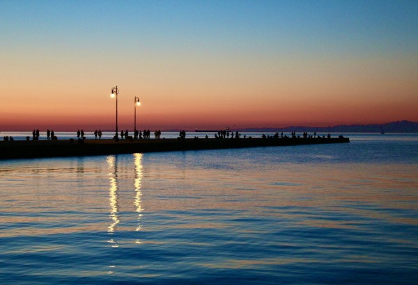 Silhouettes of people on a pier at sunset with lampposts and a reflective water surface.