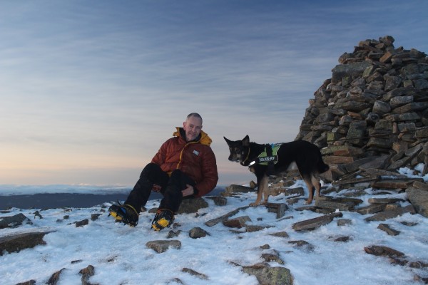 Me and Moray the dog sat next to a summit cairn. The ground is stony with a covering of hard snow. I'm wearing an old red duvet jacket, and I have crampons on my boots.