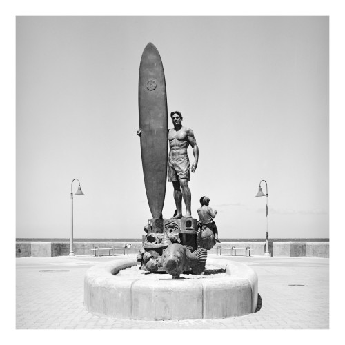 This black and white photograph features a life-sized statue of a surfer standing tall with a surfboard under his right arm. The male figure is depicted as muscular, wearing swim trunks, and looking confidently forward. Positioned on a circular base that also includes several marine-themed sculptures like sea turtles and fish, the statue celebrates coastal culture and the sport of surfing. In the background, the sky is clear, and the pavement of the promenade is patterned, suggesting this setting is along a beachfront area. Two street lamps flank the scene, enhancing its public, outdoor setting. The composition effectively conveys a tribute to surfing, capturing the spirit of the seaside activity in a communal space.