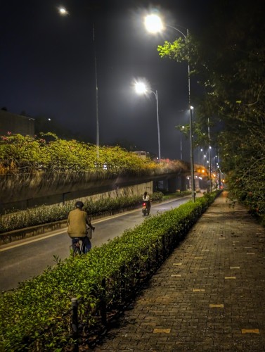 A late night photo of a long stretch of road in exurban Hanoi. We are standing on a broad, brick sidewalk that disappears into a vanishing point beyond a distant traffic light. To our left is a low hedge row separating us from the road. On said road we see a young woman on a motorbike speeding away and an elderly man on a bicycle making his slow way onward behind her. Farther left of them is the incline of an overpass that takes traffic over the approaching intersection. Flowering bushes grow from planters along its railing. Streetlights glow brightly above.