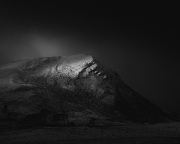 A dark black and white landscape photograph from the highlands of Scotland with rugged, snow dusted mountain emerging from the gloom and caught on fleeting sunbeam through flurries of snow. 