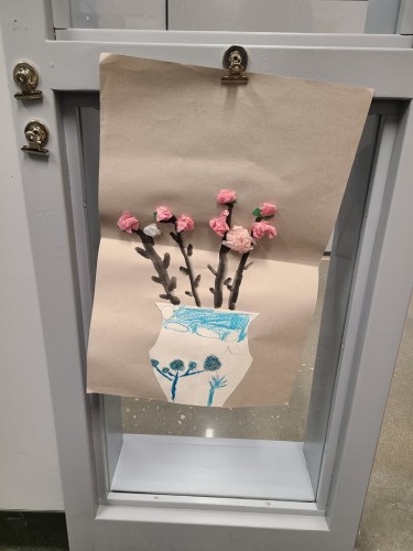 Photograph of kid's artwork. The artwork is of a white vase with blue trees / skies drawn on it. In the vase are flowers with black stems drawn on and the flowers, all shades of pink, made from crinkle paper.