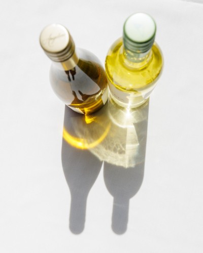 Color photo of two bottles of white wine on a white tablecloth from almost directly above. Sunlight passes through the bottles, casting both the shadows of the bottles and yellowy light filtered as it passed through the wine itself.