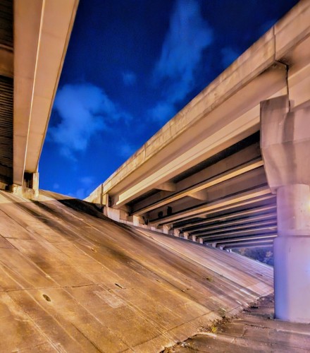 Late night view from below a large concrete highway overpass bridge. A deep blue night sky is visible between the directional roadways overhead, the dark, dirty, weathered concrete structure illuminated by underside yellow lighting. A nearby red traffic signal, out of view, reflects a pink-red shade on one of the support columns.