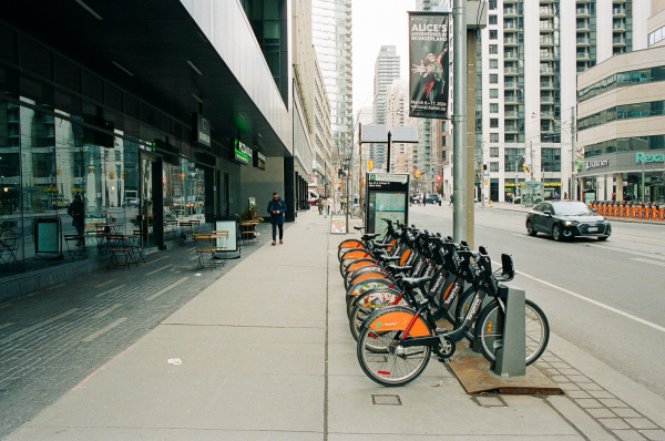 A row of black and orange festooned bicycles line the sidewalk by a downtown office building while a man walks by café seating looking at his cellphone. 