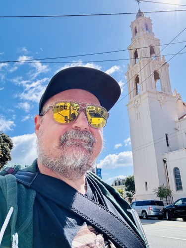 A selfie of me on a sunny day; I’m wearing mirrored gold sunglasses and Mission Dolores is behind me.