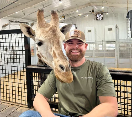 A bearded white man sits on a raised platform and smiles while the head of a giraffe rests on his shoulder.