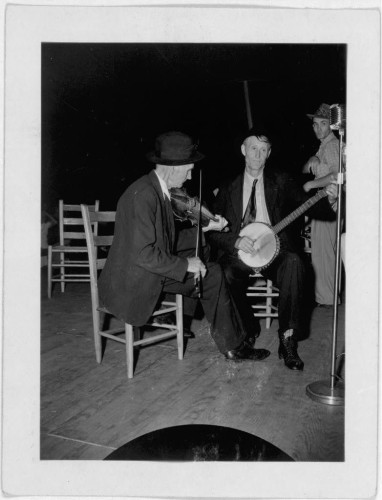  The image appears to be a vintage black and white photograph. It captures a moment of musical performance, featuring two men engrossed in playing their respective instruments: one individual is holding a fiddle while the other plays a banjo. They are positioned on a stage with wooden flooring, as indicated by the visible planks beneath their feet. In the background, there's a third person who seems to be accompanying them on guitar, given the outline of an instrument and the presence of a microphone stand. The text in the image provides context that this performance is at "Mountain Music Festival," suggesting it could be from the early 20th century when such festivals were more prevalent. However, without specific dates or additional information, it's difficult to pinpoint the exact years of the photograph's capture. The style and fashion depicted in the image further suggest a period that might fall within the early to mid-20th century. 