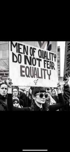 Black and white image of a lady holding up a large placard reading MEN OF QUALITY DO NOT FEAR EQUALITY