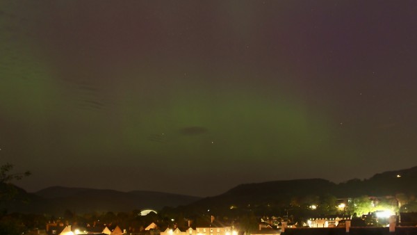 Green and purple banded areas of aurora above panoramic view above hills on the horizon and the street lights of a town below 